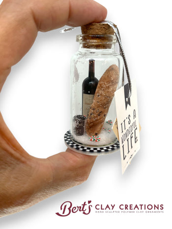 Hand holding a small glass jar with miniature bottle and bread, featuring a tag with text, on a white background. Brand 'Bert's Clay Creations' visible.